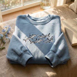 Op de afbeelding: Een lichtblauw sweatshirt met ronde hals en een geborduurd ontwerp van drie hamerhaaien die tussen kleine, gestikte gouden sterren zwemmen. Het kledingstuk heeft lange mouwen, een geribbelde kraag en een zachte, effen stoffen constructie.
