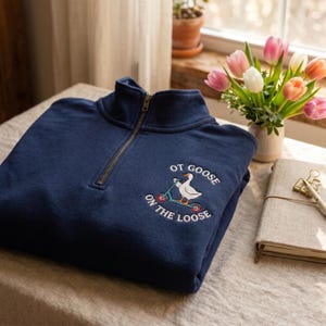 May include: A navy blue quarter-zip sweatshirt featuring an embroidered graphic of a white goose riding a light blue scooter. The text "OT GOOSE ON THE LOOSE" is arched around the goose in white lettering. The sweatshirt has a metal zipper pull.