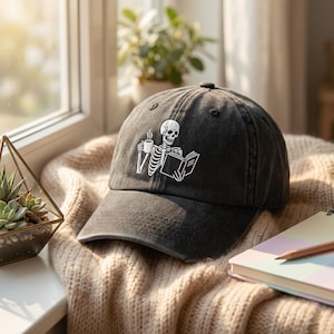 Peut inclure: Casquette de baseball noire avec un aspect vieilli. Elle présente un motif blanc d'un squelette lisant un livre et tenant une tasse de café. La casquette est posée sur une couverture en tricot beige.