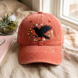 May include: A coral-coloured baseball cap with an embroidered design of a black raven holding a coffee cup, encircled by a floral wreath. The cap has a curved brim and a classic six-panel design. The background is a soft, neutral fabric.