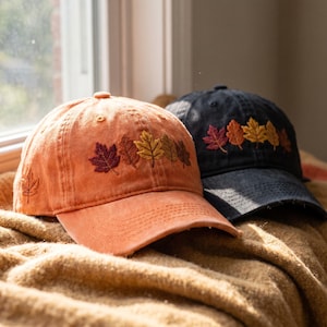 Op de afbeelding: Twee baseballpetten, één in vervaagd oranje en één in antracietzwart, elk voorzien van geborduurde herfstachtige esdoornbladmotieven in rood-, oranje- en geeltinten op de voorpanelen en een enkel blad aan de zijkant van de oranje pet.