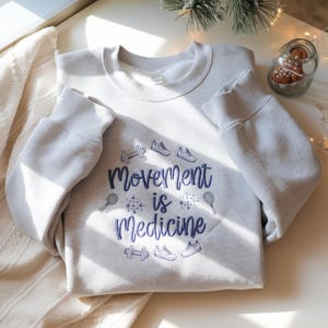May include: A light gray sweatshirt with the text "Movement is Medicine" in blue script. The design includes illustrations of dumbbells, sneakers, and tennis rackets. The sweatshirt is displayed on a white surface.