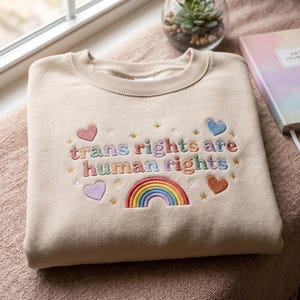 Puede incluir: Sudadera beige con bordado "trans rights are human rights", un arcoíris, corazones y estrellas. La sudadera está doblada sobre una manta rosa cerca de una ventana, con una pequeña planta y un diario en el fondo.