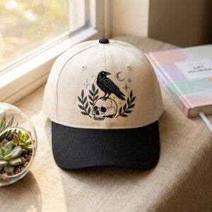 Peut inclure: Casquette de baseball beige et noire avec un motif brodé d'un corbeau noir perché sur un crâne, avec des étoiles et un croissant de lune. La casquette a une visière noire et un bouton noir sur le dessus. Le motif est entouré de feuilles.