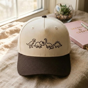 Op de afbeelding: Beige baseballpet met een bruine klep. De pet heeft een zwarte lijntekening van vier dinosaurussen op de voorkant. De pet heeft een afgeronde bovenkant en een knoop.