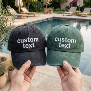 Op de afbeelding: Twee baseballpetten, één in antracietgrijs en één in bosgroen, beide met een verwassen stoffen afwerking. Elke pet is op het voorpaneel voorzien van het geborduurde opschrift custom text in witte, schreefloze hoofdletters.