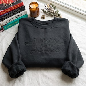 Könnte beinhalten: Ein dunkelgraues Sweatshirt mit Rundhalsausschnitt und einer schwarzen Stickerei, die ein von Blumenranken umwundenes Schwert zeigt. Unter dem Schwert ist der Schriftzug "you do not yield" in passender schwarzer Schreibschrift eingestickt.