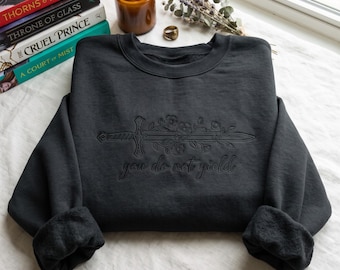 May include: A dark charcoal gray crewneck sweatshirt featuring black embroidery of a sword entwined with floral vines. Below the sword, the text "you do not yield" is embroidered in a matching black cursive script across the center chest.