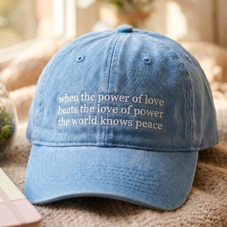 May include: A light blue denim baseball cap with a distressed finish. The front panel features white embroidered text that reads: when the power of love beats the love of power the world knows peace. The cap has a curved brim and a button top.