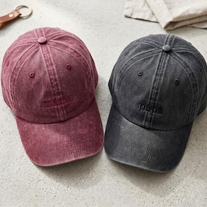 Puede incluir: Dos gorras de béisbol, una burdeos y otra gris antracita. La gorra burdeos tiene bordado "mama" en la parte delantera, y la gorra gris tiene bordado "papa" en la parte delantera. Ambas gorras tienen un aspecto vintage.