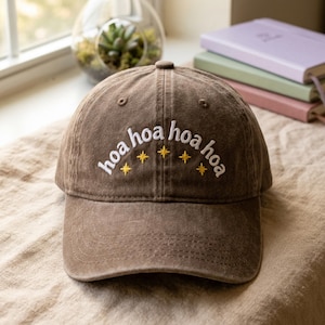 Op de afbeelding: Een bruine baseballpet met witte geborduurde tekst "hoa hoa hoa hoa" in een boogvorm, geaccentueerd door vijf kleine gele geborduurde vierpuntige sterren die direct onder de tekst op het voorpaneel van de pet zijn geplaatst.