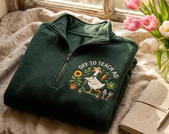 May include: A dark green quarter-zip sweatshirt featuring an embroidered graphic of a white goose wearing a hat and scarf while riding a scooter. The design includes a sunflower, carrot, watering can, and garden tools with the text OFF TO TEACH AG.