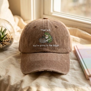 Peut inclure: Casquette de baseball marron avec l'inscription "That's it. You're going in the SOUP" et une grenouille de dessin animé. La casquette a une visière incurvée et une fermeture à sangle.