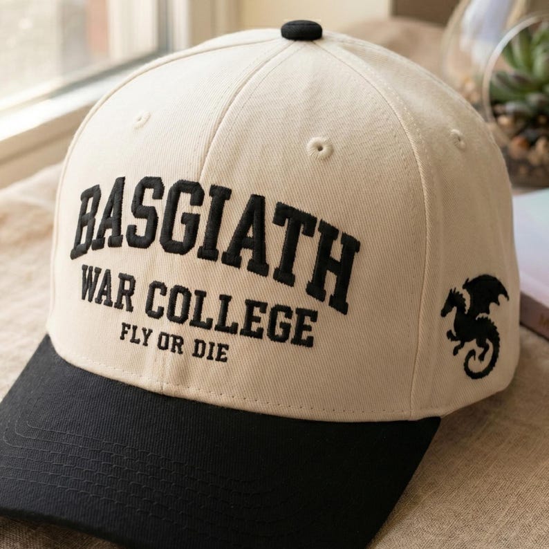 May include: Cream-colored baseball cap with a black brim and top button. The cap features the text "BASGIATH WAR COLLEGE FLY OR DIE" in black, along with a black dragon graphic on the side.