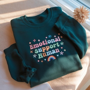 Może przedstawiać: Ciemnozielona bluza z haftowanym napisem "Emotional Support Human" w kolorowych literach, z tęczą i kwiatowymi akcentami. Bluza ma okrągły dekolt i długie rękawy.