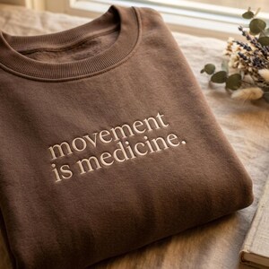 May include: A brown sweatshirt with the embroidered text "movement is medicine." The crew neck sweatshirt is folded and resting on a neutral-colored surface. Dried flowers and a book are in the background.