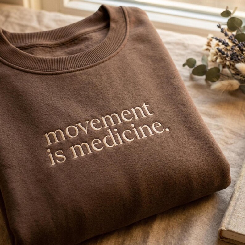 May include: A brown sweatshirt with the embroidered text "movement is medicine." The crew neck sweatshirt is folded and resting on a neutral-colored surface. Dried flowers and a book are in the background.