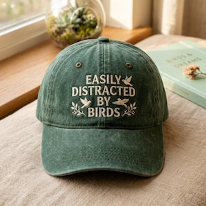 Könnte beinhalten: Grüne Baseballkappe mit dem gestickten Text "EASILY DISTRACTED BY BIRDS" in Weiß. Die Kappe hat einen gebogenen Schirm und ein klassisches Sechs-Panel-Design. Der Text wird von Vogel- und Blätterillustrationen begleitet.