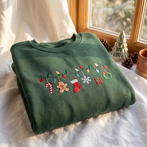 Pode incluir: Uma camisola de gola redonda verde-floresta com um fio de luzes de Natal bordado. Pendurados no fio de luzes estão um bastão de rebuçado, um boneco de gengibre, uma meia vermelha, um floco de neve branco, um presente e uma coroa.