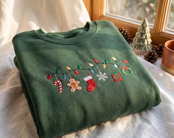 May include: A forest green crewneck sweatshirt featuring an embroidered string of colorful Christmas lights. Hanging from the light strand are a candy cane, a gingerbread man, a red stocking, a white snowflake, a gift box, and a green wreath.