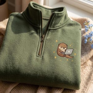 Peut inclure: Polaire vert olive avec un col montant et une fermeture &eacute;clair quart de zip. Il pr&eacute;sente une broderie d'une loutre de dessin anim&eacute; utilisant un ordinateur portable, entour&eacute;e de petites &eacute;toiles jaunes. Le tissu polaire est pli&eacute; sur une couverture en tricot beige.