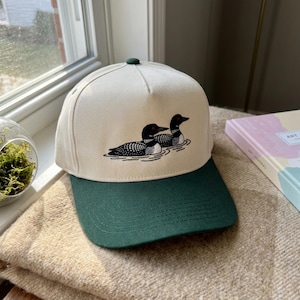 Puede incluir: Gorra de béisbol color crema con visera y botón verde oscuro. Presenta una ilustración en blanco y negro de dos somormujos nadando en el agua. Un accesorio de moda para actividades al aire libre.