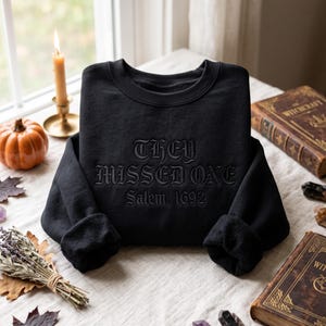 Op de afbeelding: Een zwart sweatshirt met ronde hals en ton-sur-ton zwarte borduursels op de borst. De tekst "THEY MISSED ONE" staat in een vet gotisch lettertype, met "Salem, 1692" eronder geborduurd in een kleiner, bijpassend zwart gotisch schrift.
