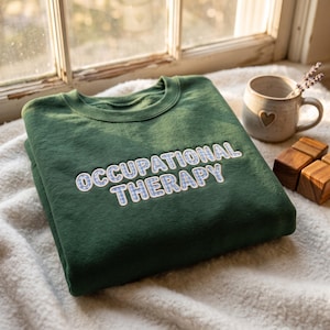 May include: A dark green crewneck sweatshirt featuring the text OCCUPATIONAL THERAPY embroidered in a light blue and white gingham pattern. The lettering is outlined in white thread, creating a bold and clear design on the front of the garment.