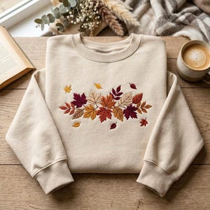 May include: A beige crewneck sweatshirt featuring an embroidered cluster of autumn leaves in shades of burgundy, burnt orange, golden yellow, and tan. The design is centered on the chest, showcasing detailed leaf veins and varied foliage shapes.