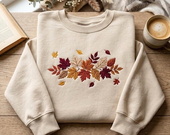 May include: A beige crewneck sweatshirt featuring an embroidered cluster of autumn leaves in shades of burgundy, burnt orange, golden yellow, and tan. The design is centred on the chest, showcasing detailed leaf veins and varied foliage shapes.