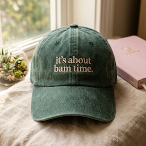 May include: A dark green baseball cap with the embroidered text "it's about bam time." The hat is made of a soft, textured fabric and has a curved brim. A pink journal is visible in the background.