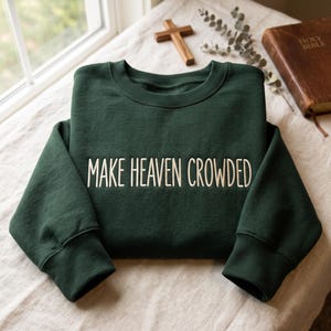 Puede incluir: Una sudadera de cuello redondo en color verde bosque oscuro con el texto "MAKE HEAVEN CROWDED" bordado en hilo de color crema claro en el pecho. La prenda tiene mangas largas con puños acanalados y un cuello acanalado.