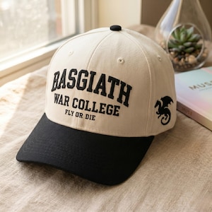 May include: A beige baseball cap with a black brim. The cap features the text "BASGIATH WAR COLLEGE FLY OR DIE" in black, along with a black dragon graphic on the side. The cap has a black button on top.