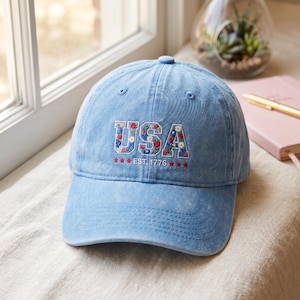 May include: A light blue acid-wash baseball cap featuring the embroidered text USA in a block font filled with small red, white, and blue floral patterns. Below the text, the words EST. 1776 are flanked by three small red stars on each side.