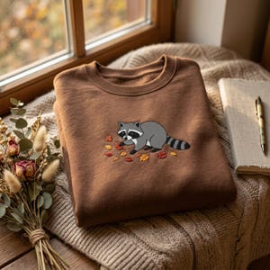 May include: A brown crewneck sweatshirt featuring an embroidered graphic of a grey raccoon with a striped tail, positioned among small, colorful autumn leaves in shades of orange, yellow, and red, centered on the chest of the garment.