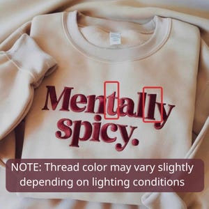 May include: A beige crewneck sweatshirt featuring the embroidered text Mentally spicy. in a dark red thread. The design includes two red rectangular outlines highlighting specific letters within the word Mentally. The fabric has a smooth texture.