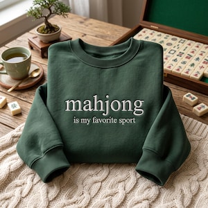 Op de afbeelding: Een donkergroene sweater met ronde hals en witte geborduurde tekst "mahjong is my favorite sport" in het midden op de borst. Het kledingstuk heeft lange mouwen met geribbelde boorden en een geribbelde halslijn, gemaakt van effen stof.