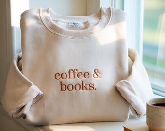 May include: A beige sweatshirt with the embroidered text "coffee & books." The sweatshirt is folded and resting on a windowsill, with a cup of coffee and a stack of books nearby. The sweatshirt has a crew neck and long sleeves.