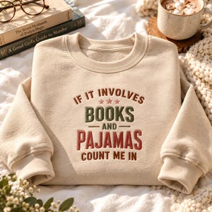 May include: A beige crewneck sweatshirt featuring embroidered text that reads IF IT INVOLVES BOOKS AND PAJAMAS COUNT ME IN, with three small stars above the word BOOKS. The text is rendered in a combination of brown, green, and red lettering.
