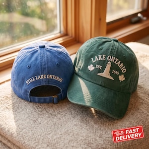 Puede incluir: Una gorra de béisbol azul con el texto STILL LAKE ONTARIO bordado en la parte posterior y una gorra de béisbol verde con el texto LAKE ONTARIO EST. 1615, una ilustración de faro y dos iconos de hoja de arce bordados en el frente.