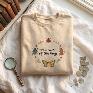 May include: A beige crewneck sweatshirt featuring a circular graphic of various insects including a ladybug, a beetle, and a butterfly, surrounding the text "the last of the bugs" written in a dark script font with small green leaf accents.
