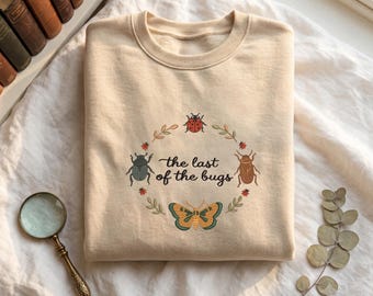 Puede incluir: Una sudadera de cuello redondo en color beige con un gráfico circular de varios insectos, incluyendo una mariquita, un escarabajo y una mariposa, rodeando el texto "the last of the bugs" en letra cursiva oscura con hojas verdes.