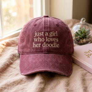 Könnte beinhalten: Burgunderfarbene Baseballkappe mit dem gestickten Text "just a girl who loves her doodle". Die Kappe ist aus einem weichen, strukturierten Stoff gefertigt und hat einen gebogenen Schirm. Der Hut liegt auf einer beigen Oberfläche.