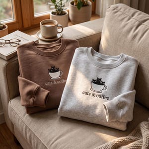 Op de afbeelding: Twee sweaters met ronde hals, één in effen bruin en één in gemêleerd grijs, elk voorzien van een borduursel van drie zwarte katten in een witte koffiekop met de tekst "cats & coffee" gestikt in donkerbruin garen onder het ontwerp.
