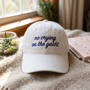 May include: A white baseball cap featuring the embroidered text "no crying on the yacht" in dark blue cursive lettering. The cap has a structured crown, a curved brim, and a button at the top center, showcasing a clean and simple design.