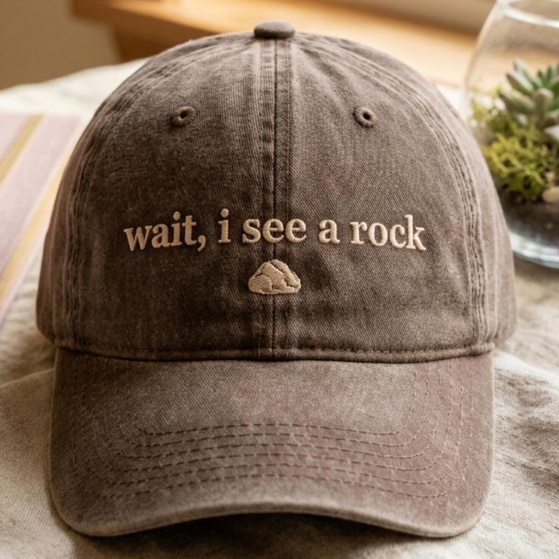 Puede incluir: Gorra de b&eacute;isbol marr&oacute;n con la frase bordada "wait, i see a rock" en color beige claro. Un peque&ntilde;o gr&aacute;fico de roca est&aacute; bordado debajo del texto. La gorra tiene una visera curva y un bot&oacute;n en la parte superior.