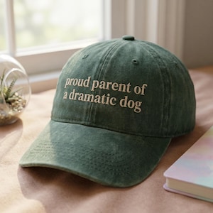 May include: A green baseball cap with the embroidered text "proud parent of a dramatic dog". The hat has a curved brim and a rounded top. The hat is sitting on a brown surface.