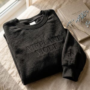 May include: Black sweatshirt with the words "AIRPLANE MODE" embroidered across the chest. A small airplane is embroidered on the left sleeve. A book titled "TRAVEL NOTES" and some blue flowers are in the background.