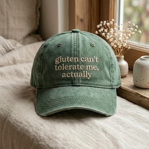 May include: A green baseball cap with the embroidered text "gluten can't tolerate me, actually". The cap has a curved brim and a classic six-panel design. The hat is made of a soft, worn material.