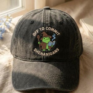 K&ouml;nnte beinhalten: Dunkelgraue Baseballkappe mit dem gestickten Schriftzug "OFF TO COMMIT SHENANIGANS". Die Kappe zeigt einen Cartoon-Frosch mit Zaubererhut und -robe, der einen Stab und ein Buch h&auml;lt. Der Frosch ist gr&uuml;n, der Hut braun und der Text wei&szlig;.